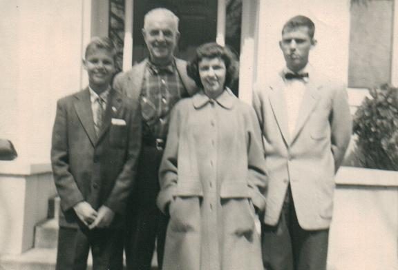 A black-and-white snapshot of four people on the steps of a white clapboard porch: a boy of about fourteen in a jacket and tie at the left, a white-haired man in a checked shirt and dark jacket behind him, a dark-haired woman in a pale belted coat in front, and a young man in a light jacket and bow tie at the right.