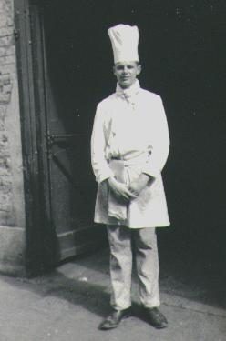A black-and-white snapshot of a young man standing in a stone doorway in a cook's white jacket and long apron and a tall white chef's toque, hands folded in front of him.