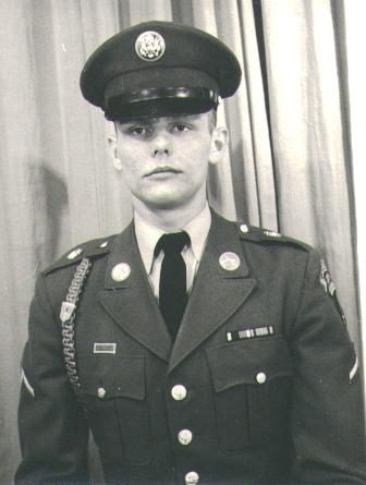 A formal black-and-white head-and-shoulders portrait of a young soldier in US Army dress greens, service cap squared, dark tie, ribbon bar and unit insignia on the lapels, a braided cord over the left shoulder, looking straight at the camera without expression.