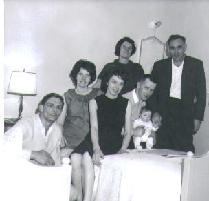 A black-and-white snapshot of seven people crowded onto and around a bed in a small apartment: a young man in shirtsleeves sitting on the bed's edge at the left, two women beside him, a third standing behind, a fourth holding a baby upright in the middle, and a man in a shirt and tie standing at the right. A lamp and a headboard behind them.