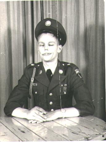 A black-and-white photograph of a young soldier in US Army dress greens and a service cap, seated at a plain wooden table in front of a curtain, hands folded on the table, eyes shut and grinning broadly. A shoulder patch and a braided cord are visible on the left shoulder.