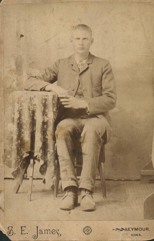 A sepia cabinet-card portrait of a young man of about twenty, seated beside a draped table, one arm resting on it, in a light jacket and trousers and heavy work shoes. The card's mount is printed 'S. E. James' at the left and 'SEYMOUR, IOWA.' at the right.