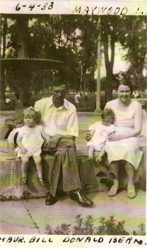 A hand-tinted snapshot of four people on a low park wall among trees: a small girl in a white dress at the left, a man in a pale shirt and dark trousers seated in the middle, a baby in white on a woman's lap, and a young woman in a pale dress at the right. Pencilled along the top edge: '6-4-33' and 'MAYWOOD IL'. Along the bottom: 'MAUR. BILL DONALD BERN.'
