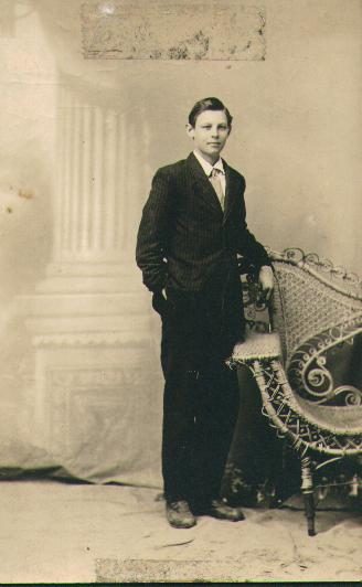 A full-length studio portrait of a boy of about fourteen in a dark striped suit and a light tie, hands in his pockets, standing beside a wicker armchair in front of a painted backdrop of a stone column and drapery.