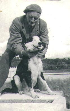 A black-and-white snapshot of an elderly man in a flat cap and tweed jacket crouching on a stone step, both hands on a black-and-white collie-type dog standing up against him; a white gate and open country behind.