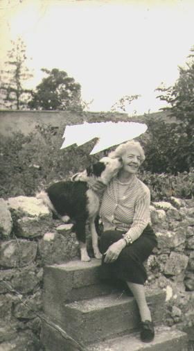 A black-and-white snapshot of an elderly woman in a light cardigan and dark skirt sitting on stone steps in a rockery garden, one hand on the back of a black-and-white collie-type dog standing beside her; a stone wall and trees behind. A pale scratch crosses the print above her head.