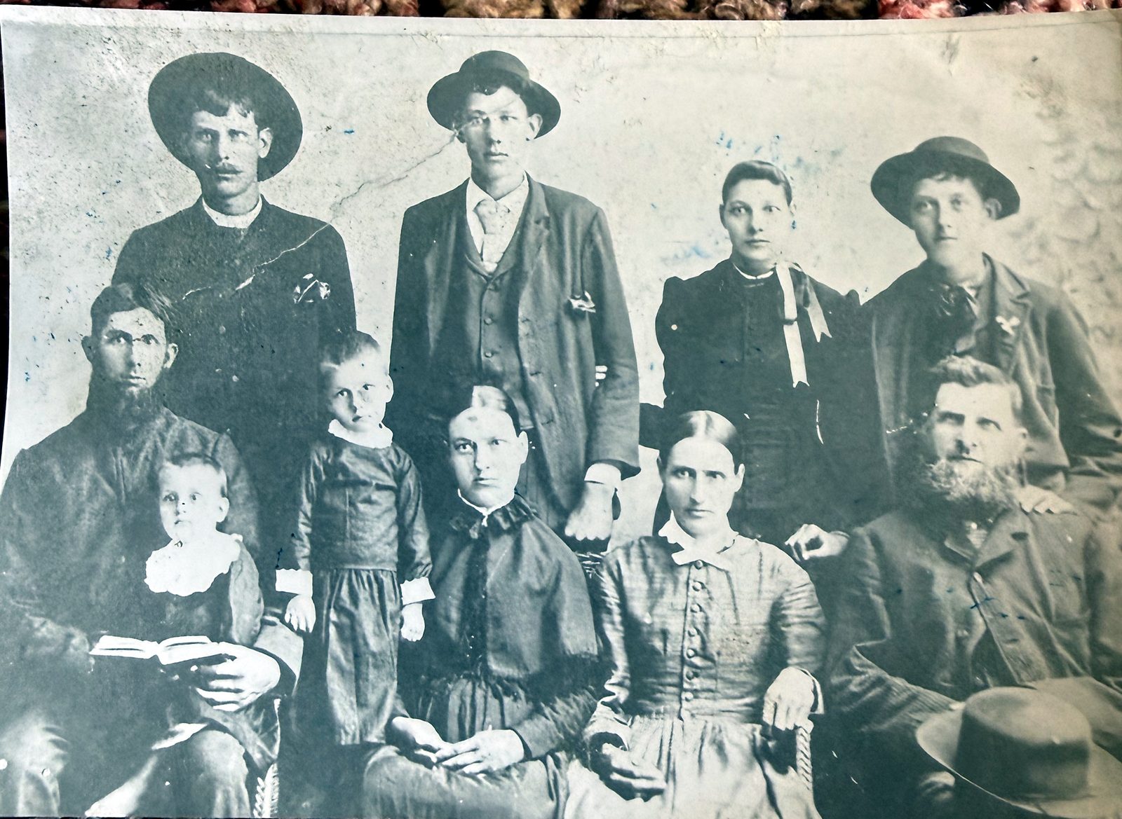 A studio group portrait of ten people, about 1890. Four adults stand across the back — three young men in hats and a young woman with a light bow at her collar. Six sit in front: a bearded man in glasses with a baby on his knee, a small boy standing at his side, two women, and at the right a white-bearded older man with his hat resting on his knee. The print is creased and spotted.