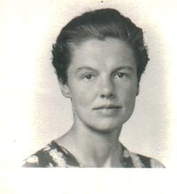 A small black-and-white head-and-shoulders portrait of a young woman with dark hair swept back, in a patterned dress with a thin chain at her neck, looking straight at the camera against a plain pale background.