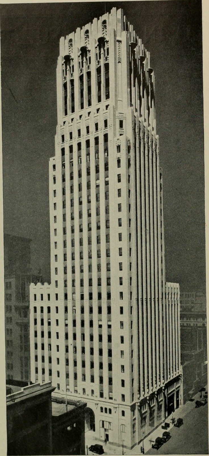 A 1930 photograph of a tall Art Deco office tower seen from slightly above street level. Its pale terracotta front rises in stepped setbacks to a crown of fluted piers and ornamental lanterns; lower buildings stand around it and period motor cars are parked in the street below.