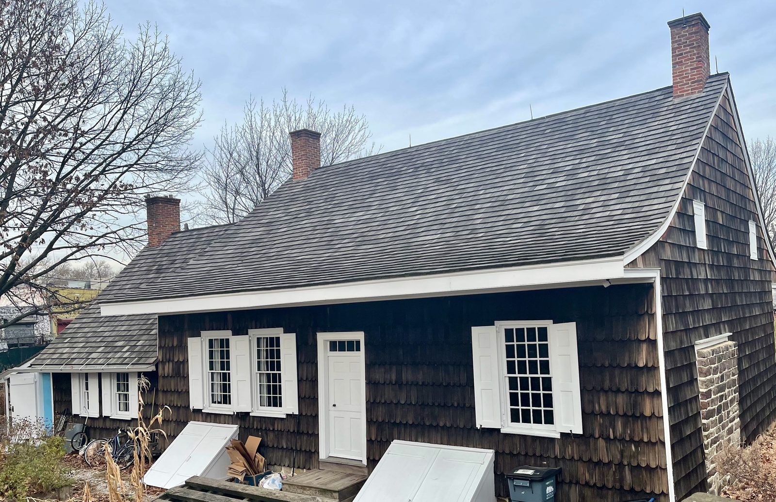 A low wooden farmhouse photographed on a grey winter day. Its long shingled roof sweeps down in a curve over the front wall and out to a shallow eave, the walls are clad in weathered dark shingles, and there are three brick chimneys. White-painted sash windows with shutters and a plain white door face the yard; a lower wing extends to the left. Bare trees, dead leaves and a modern city fence stand behind.